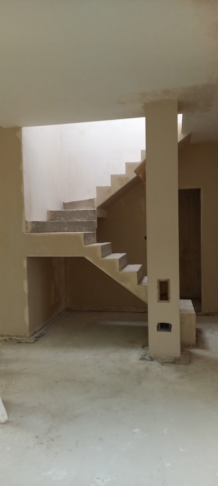 Wnętrze w trakcie tynkowania maszynowego, widoczne schody, kolumna z przygotowanymi otworami na instalacje elektryczne i surowy tynk na ścianach.