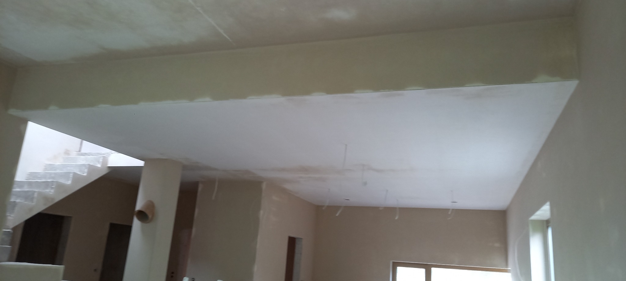 Wnętrze w stanie deweloperskim po nałożeniu jasnych tynków na ścianach i suficie, widoczne schody w tle.