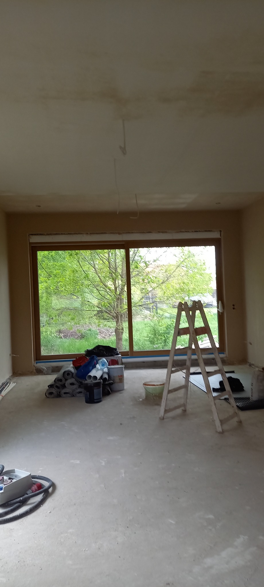 Pokój w trakcie prac wykończeniowych, widoczne świeżo położone tynki na ścianach, okno z widokiem na zieleń, drabina malarska i materiały budowlane porozrzucane po podłodze.