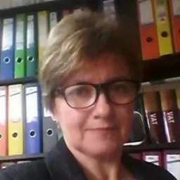 Portret kobiety w średnim wieku z krótkimi, kręconymi włosami, ubranej w ciemną marynarkę, z uśmiechem na twarzy, na tle regału z segregatorami i teczkami, w tym segregatorów z napisem VAT.