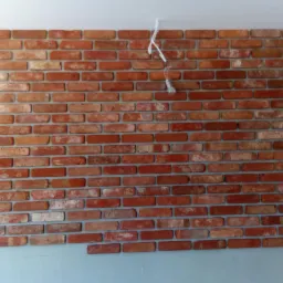 Ściana z czerwonej cegły dekoracyjnej w trakcie montażu w Rumii, widoczne wiszące przewody oświetleniowe.