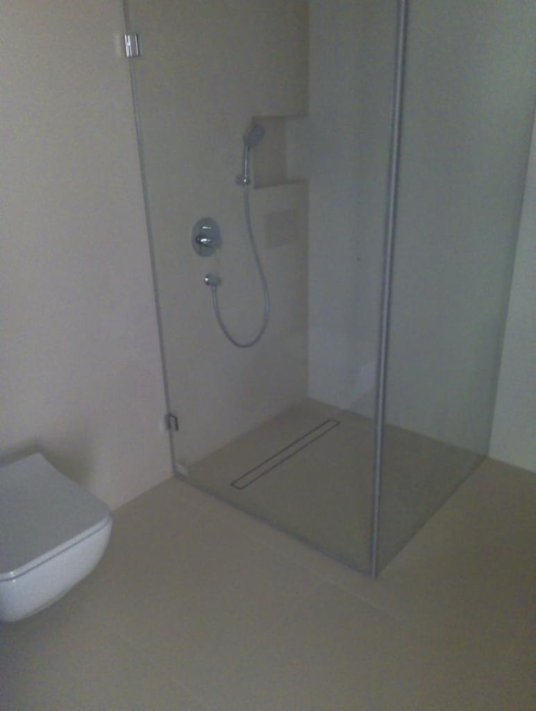 Nowoczesna kabina prysznicowa typu walk-in z odpływem liniowym w podłodze, widoczna toaleta z deską sedesową wolnoopadającą, ściany wykończone jasnymi płytkami ceramicznymi.