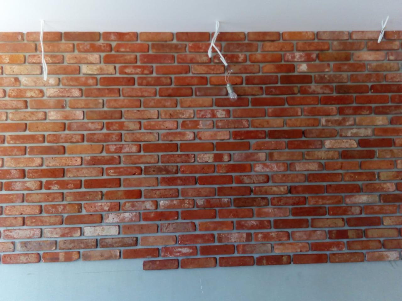 Ściana z czerwonej cegły dekoracyjnej w trakcie montażu w Rumii, widoczne wiszące przewody oświetleniowe.