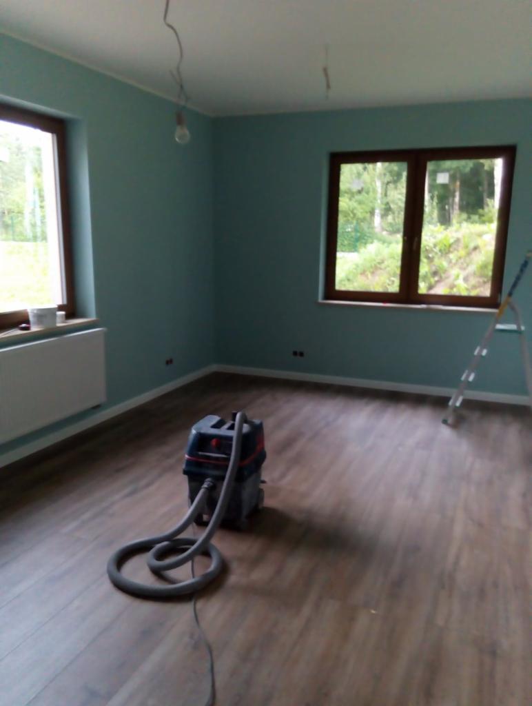 Pokój po malowaniu ścian na turkusowo, z drewnopodobną podłogą, odkurzaczem budowlanym na środku i drabiną opartą o ścianę przy oknie.