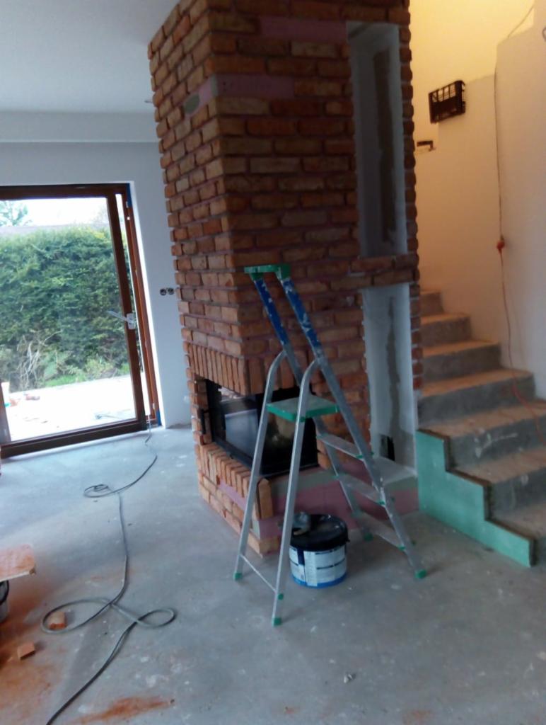 Murowany kominek z cegły w trakcie budowy, widoczna drabina i puszka farby, surowe schody w tle, wnętrze w stanie deweloperskim.