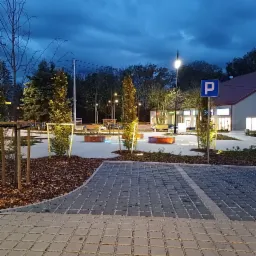 Nowo zaaranżowana przestrzeń miejska o zmierzchu, z młodymi drzewami zabezpieczonymi drewnianymi konstrukcjami, nowoczesnymi ławkami z podświetleniem LED, oświetleniem parkowym i brukowanym...