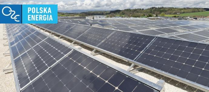 Rozległa instalacja paneli słonecznych na płaskim dachu budynku, z logo 'Polska Energia' w lewym górnym rogu i zielonym krajobrazem w tle.