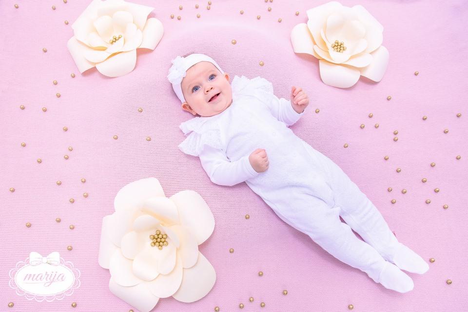 Uśmiechnięte niemowlę w białym kombinezonie z falbankami i opasce, leżące na różowym tle z dekoracyjnymi kwiatami i złotymi koralikami.