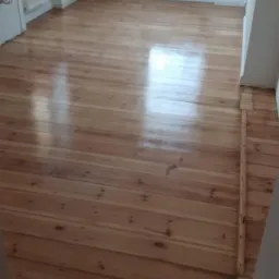 Wycyklinowana podłoga z desek w jasnym odcieniu drewna, widoczne ślady po cyklinowaniu, fragment grzejnika pod oknem i białe ściany w tle.