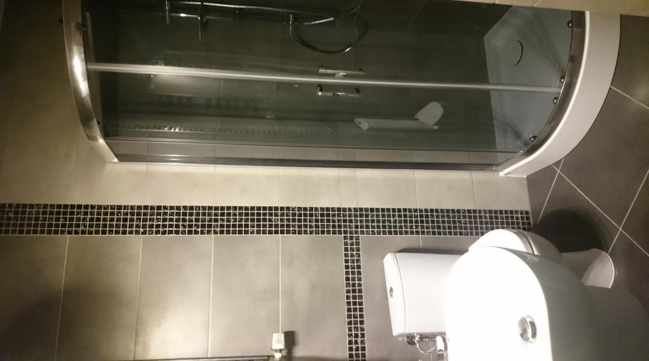 Zamontowana kabina prysznicowa z przesuwnymi drzwiami i brodzikiem, widoczna toaleta, ściany wyłożone szarymi płytkami z dekoracyjnym pasem mozaiki w kolorze czarnym i srebrnym.