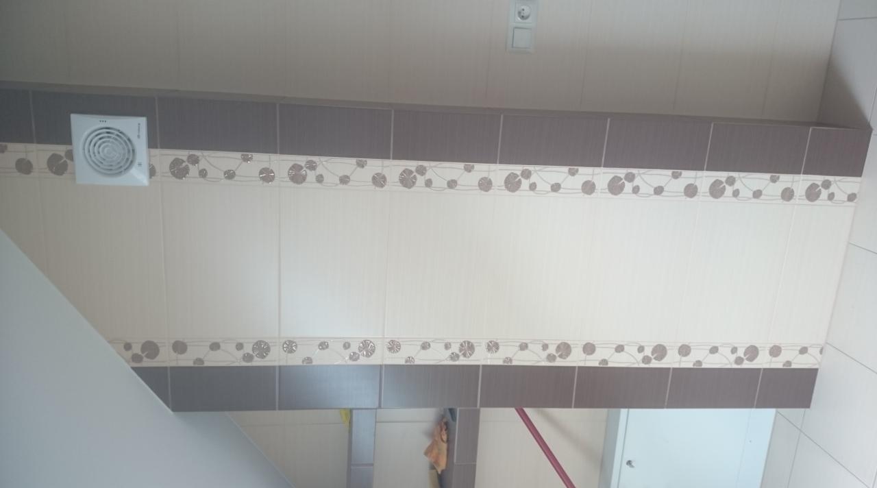 Wykończona ściana łazienki z beżowymi i brązowymi płytkami, dekoracyjnym pasem z motywem kwiatowym i wentylatorem w rogu.