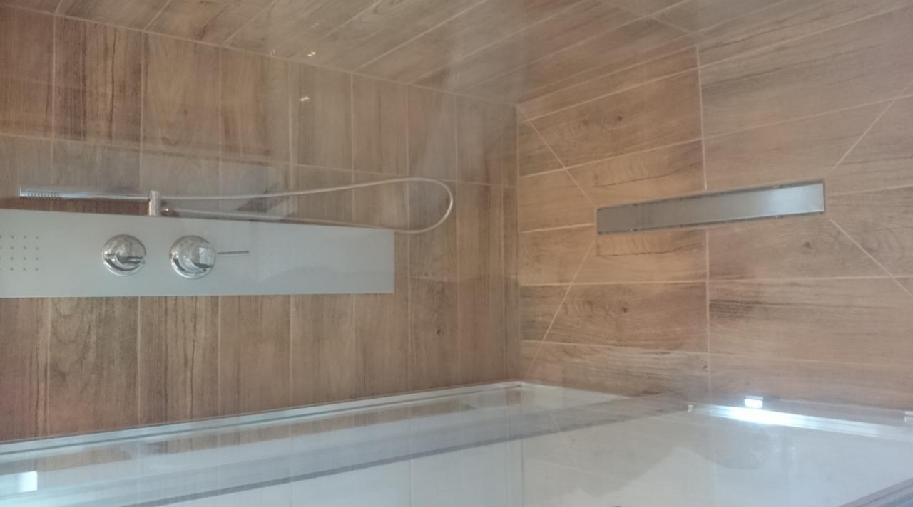 Kabina prysznicowa wyłożona płytkami imitującymi drewno z panelem prysznicowym i odpływem liniowym w podłodze.