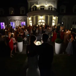 Para młoda stoi tyłem, otoczona gośćmi z zimnymi ogniami przed oświetlonym budynkiem w nocy.