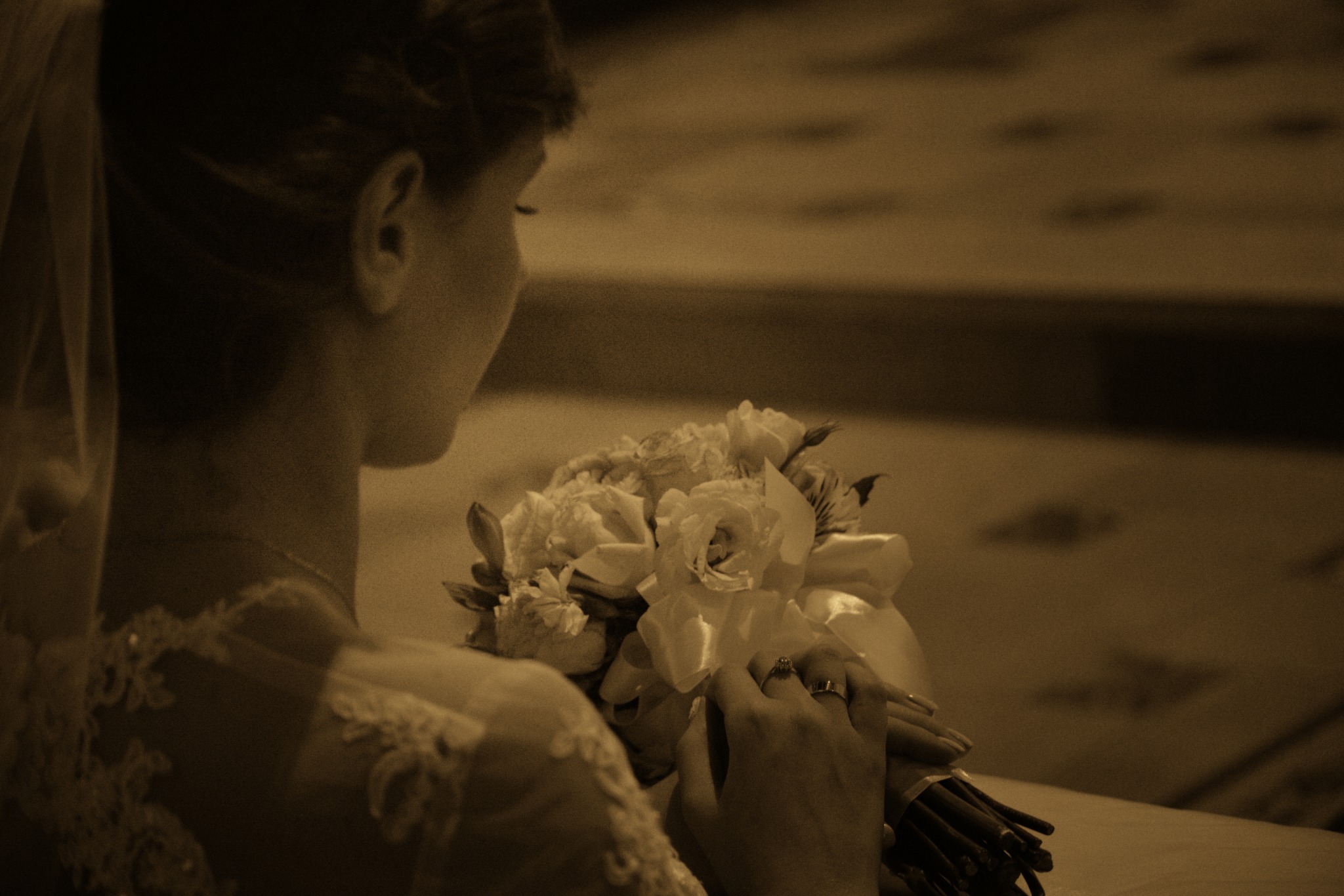 Panna młoda w sukni z koronkowymi zdobieniami trzyma bukiet białych róż, widoczny pierścionek zaręczynowy, zdjęcie utrzymane w sepii.