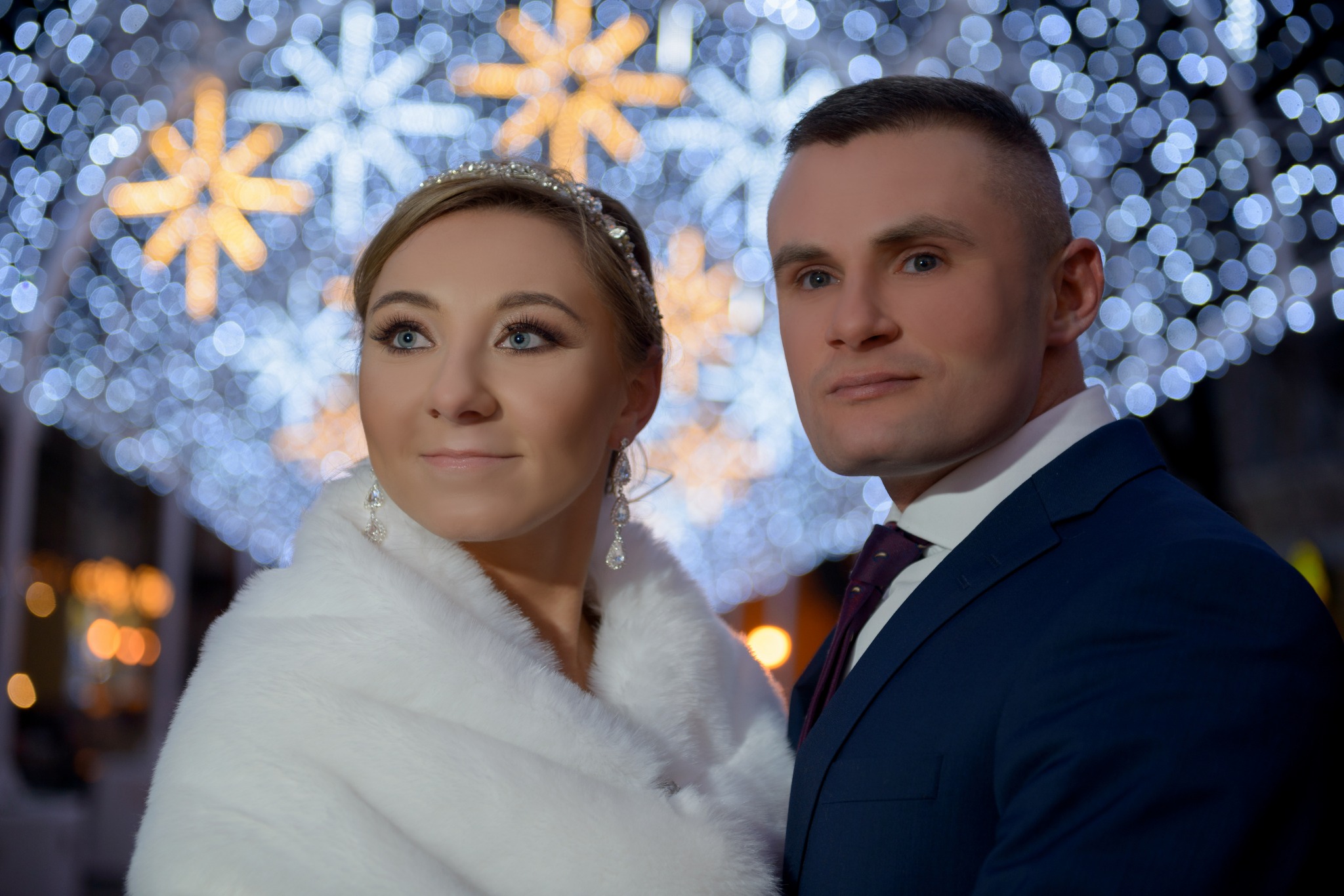 Portret pary młodej: kobieta w białym futrze i diademie, mężczyzna w granatowym garniturze i bordowym krawacie, na tle rozmytych światełek w kształcie gwiazd.