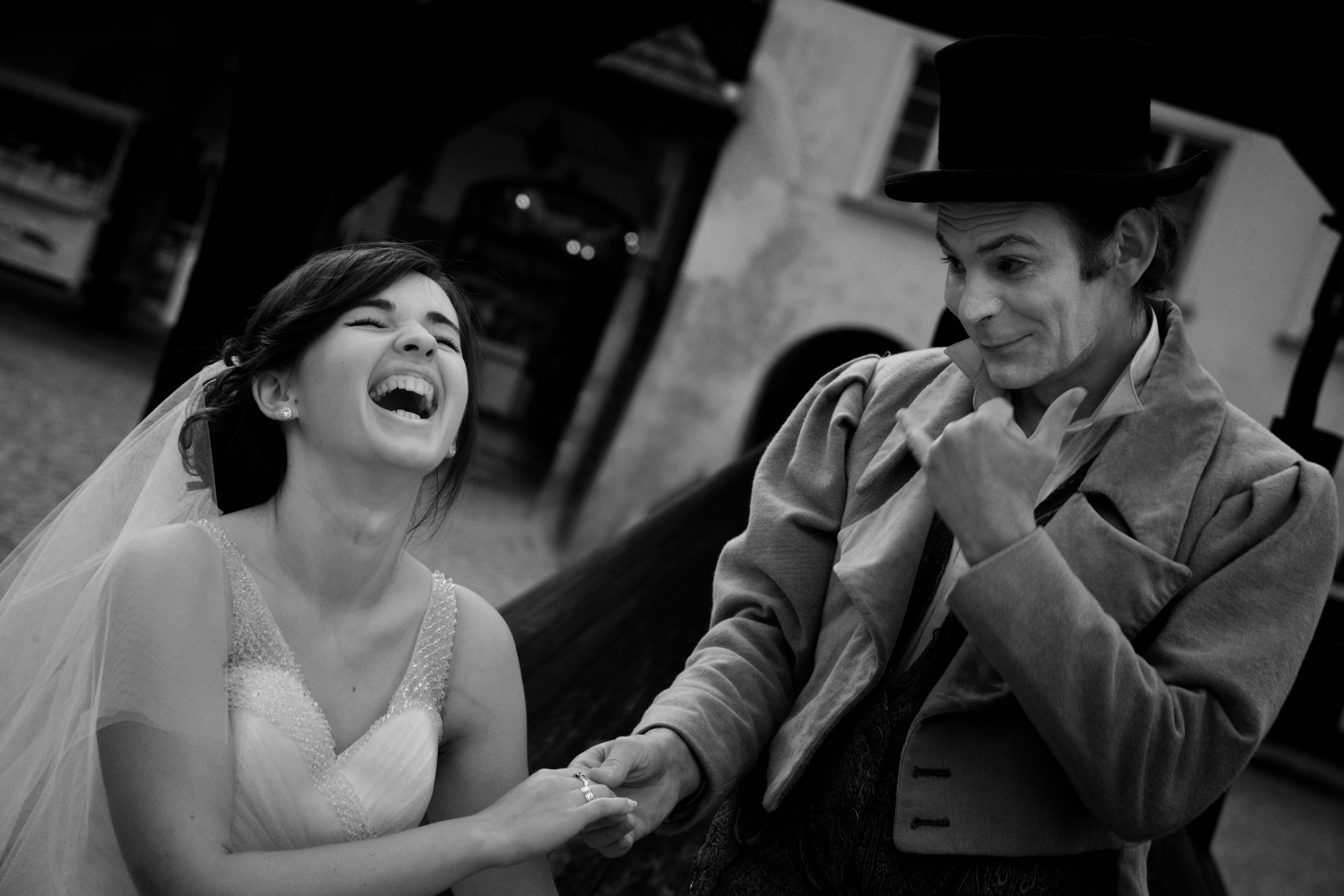 Czarno-białe zdjęcie panny młodej w sukni z kryształkami, śmiejącej się z klauna w cylindrze i fraku, trzymających się za ręce na tle budynku.