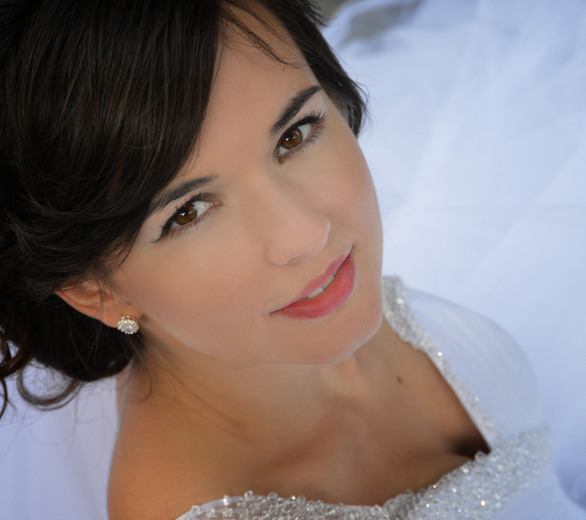 Portret uśmiechniętej panny młodej z brązowymi oczami, w białej sukni z kryształkami, z delikatnym makijażem i błyszczącymi kolczykami, sfotografowana z dołu.