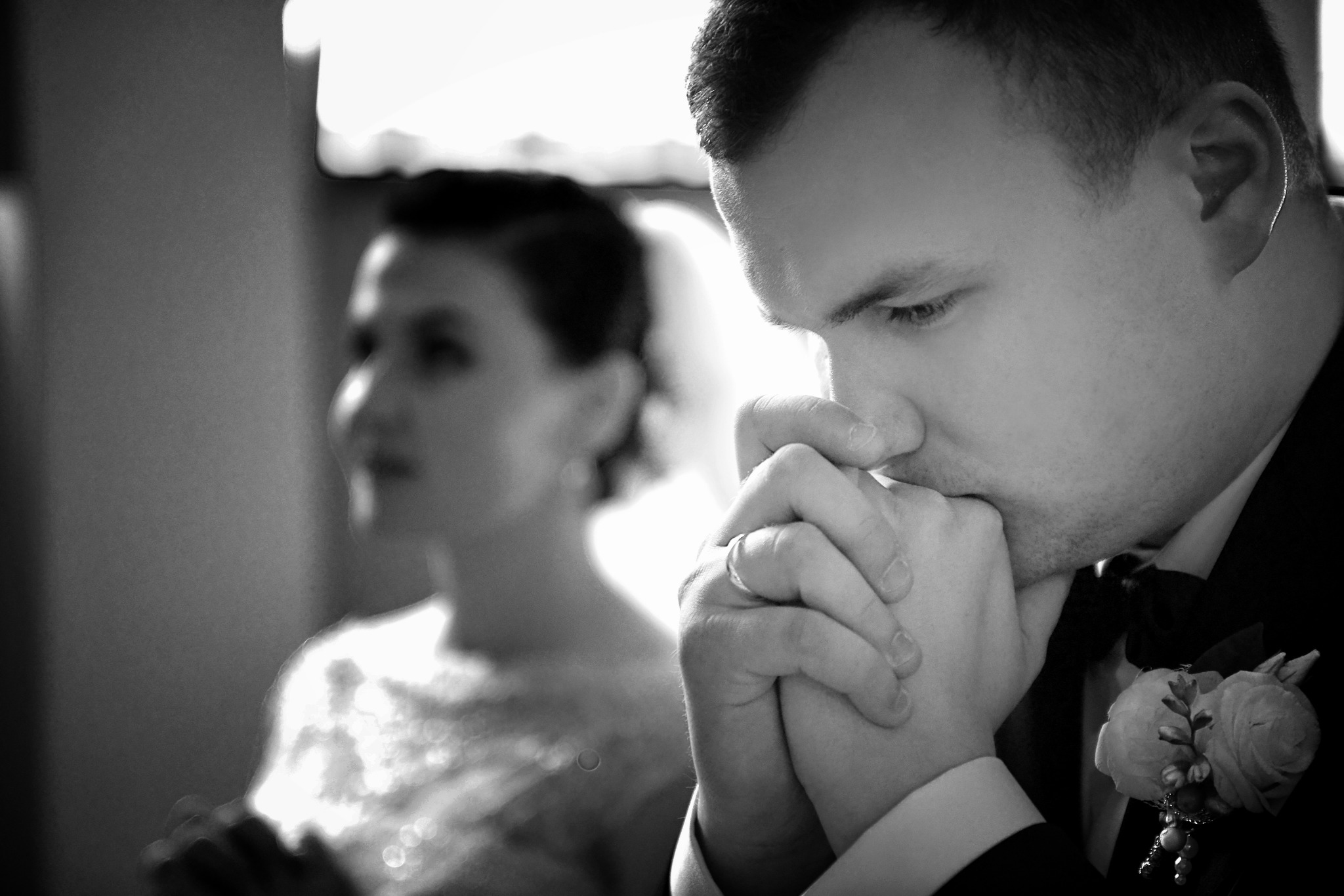 Czarno-białe zdjęcie pana młodego w skupieniu, z dłońmi złożonymi przy ustach i butonierką z jasnych róż, panna młoda w tle poza ostrością.