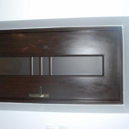 Kryształ - Ciemnobrązowe drzwi wewnętrzne z poziomym przeszkleniem i mosiężnym haczykiem na ścianie pomalowanej na biało z widocznymi gniazdkami elektrycznymi poniżej.