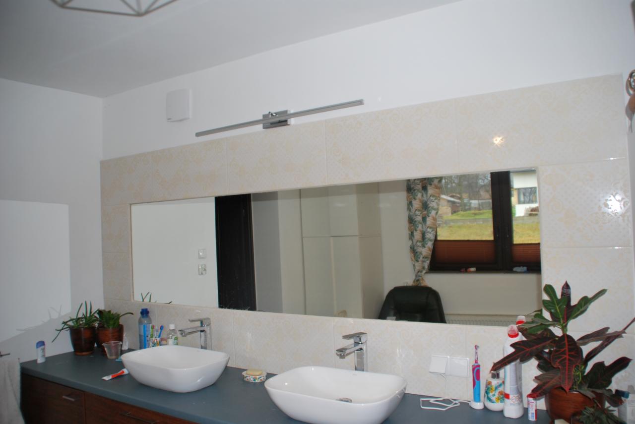 Umywalki nablatowe w łazience po remoncie, obudowane płytkami z delikatnym wzorem, z dużym lustrem w tle widoczne okno z roletami i rośliny doniczkowe.