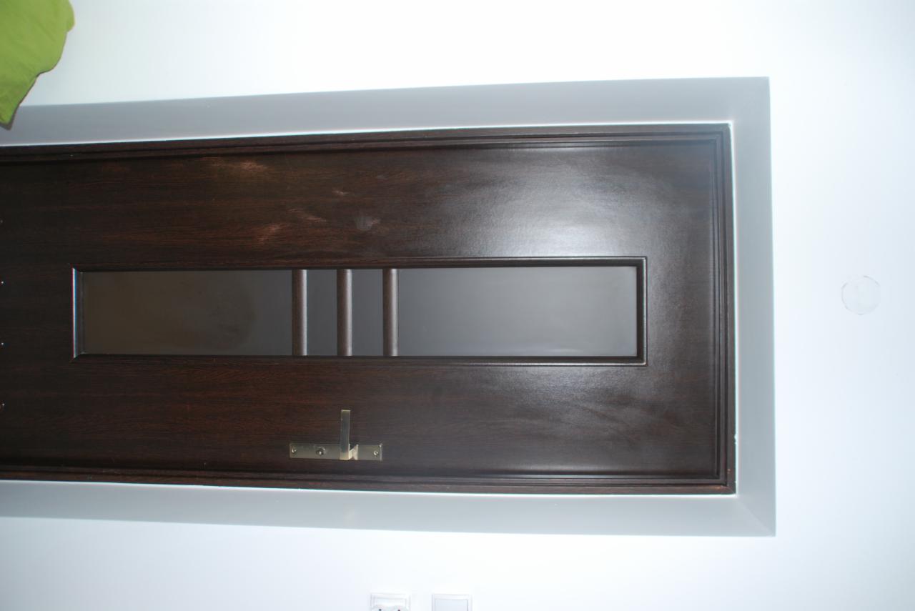 Ciemnobrązowe drzwi wewnętrzne z poziomym przeszkleniem i mosiężnym haczykiem na ścianie pomalowanej na biało z widocznymi gniazdkami elektrycznymi poniżej.