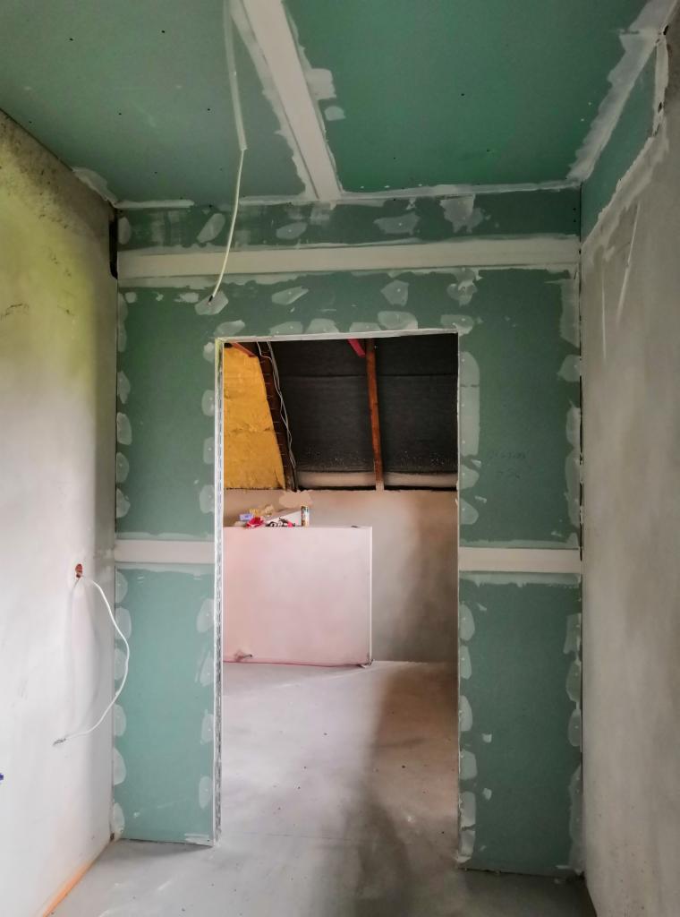 Wnętrze w trakcie prac wykończeniowych, widoczne zielone płyty na ścianach i suficie, łączenia wzmocnione taśmą, surowy stan, widok na pomieszczenie obok z izolacją i elementami konstrukcyjnymi dachu.