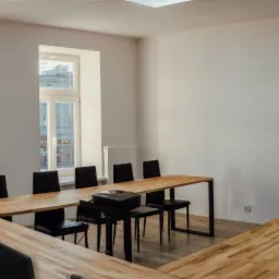 Jasne, przestronne biuro z dużym stołem konferencyjnym w kształcie litery U, czarnymi krzesłami, flipchartem i oknami z widokiem na budynki miejskie.