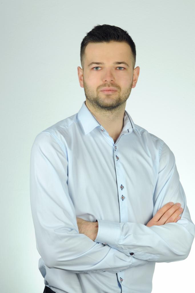 Portret mężczyzny w jasnoniebieskiej koszuli z rękami skrzyżowanymi na piersiach, stojącego na jasnym tle.