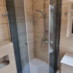 Kabina prysznicowa narożna z ciemnym szkłem, zamontowana w wykończonej łazience z beżowymi płytkami imitującymi drewno i białym wyposażeniem.