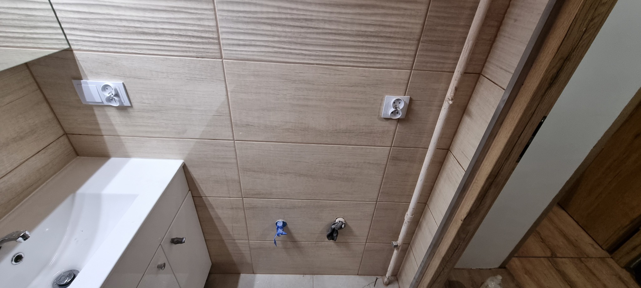 Jasne, drewnopodobne płytki ścienne w łazience, z białym zlewem, widoczne gniazdka elektryczne i wyprowadzenia hydrauliczne.