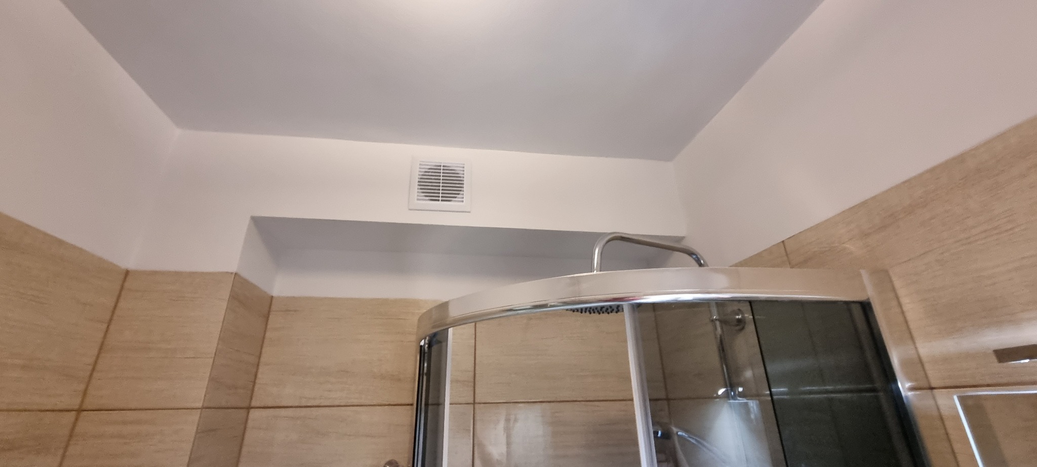 Wykończona łazienka z kabiną prysznicową z chromowanymi detalami i beżowymi płytkami ceramicznymi na ścianach, widoczny wentylator sufitowy.