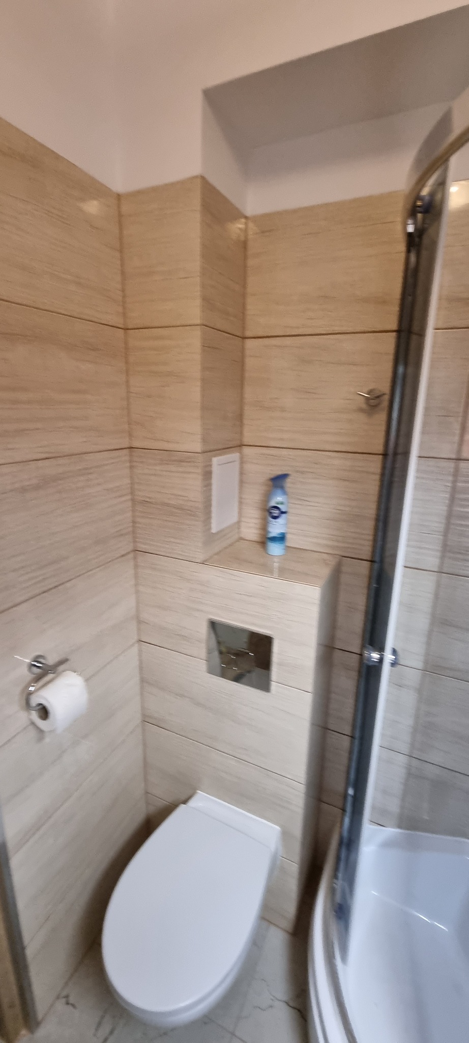 Nowoczesna łazienka z beżowymi płytkami imitującymi drewno, wisząca toaleta, chromowany przycisk spłuczki i niebieski odświeżacz powietrza na półce, kabina prysznicowa z widoczną ramą.