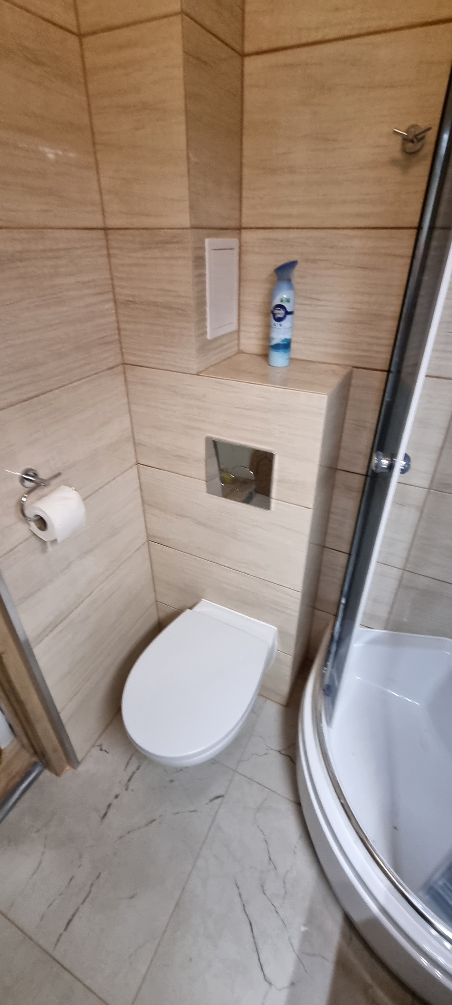 Wykończona łazienka z beżowymi płytkami imitującymi drewno, wiszącą toaletą z chromowanym przyciskiem spłukiwania, kabiną prysznicową z białym brodzikiem i odświeżaczem powietrza.