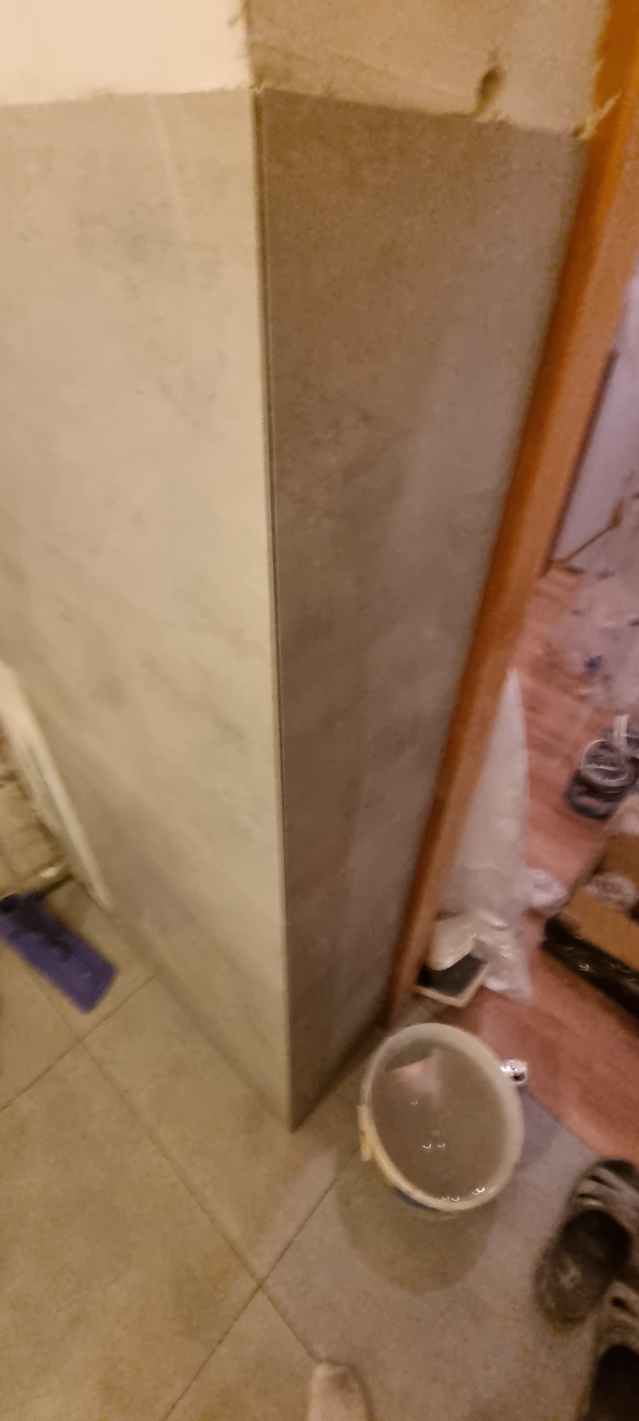Świeżo położone płytki ścienne w odcieniach szarości i beżu, widoczny fragment podłogi z płytek, wiadro z wodą i narzędzia glazurnicze w tle.