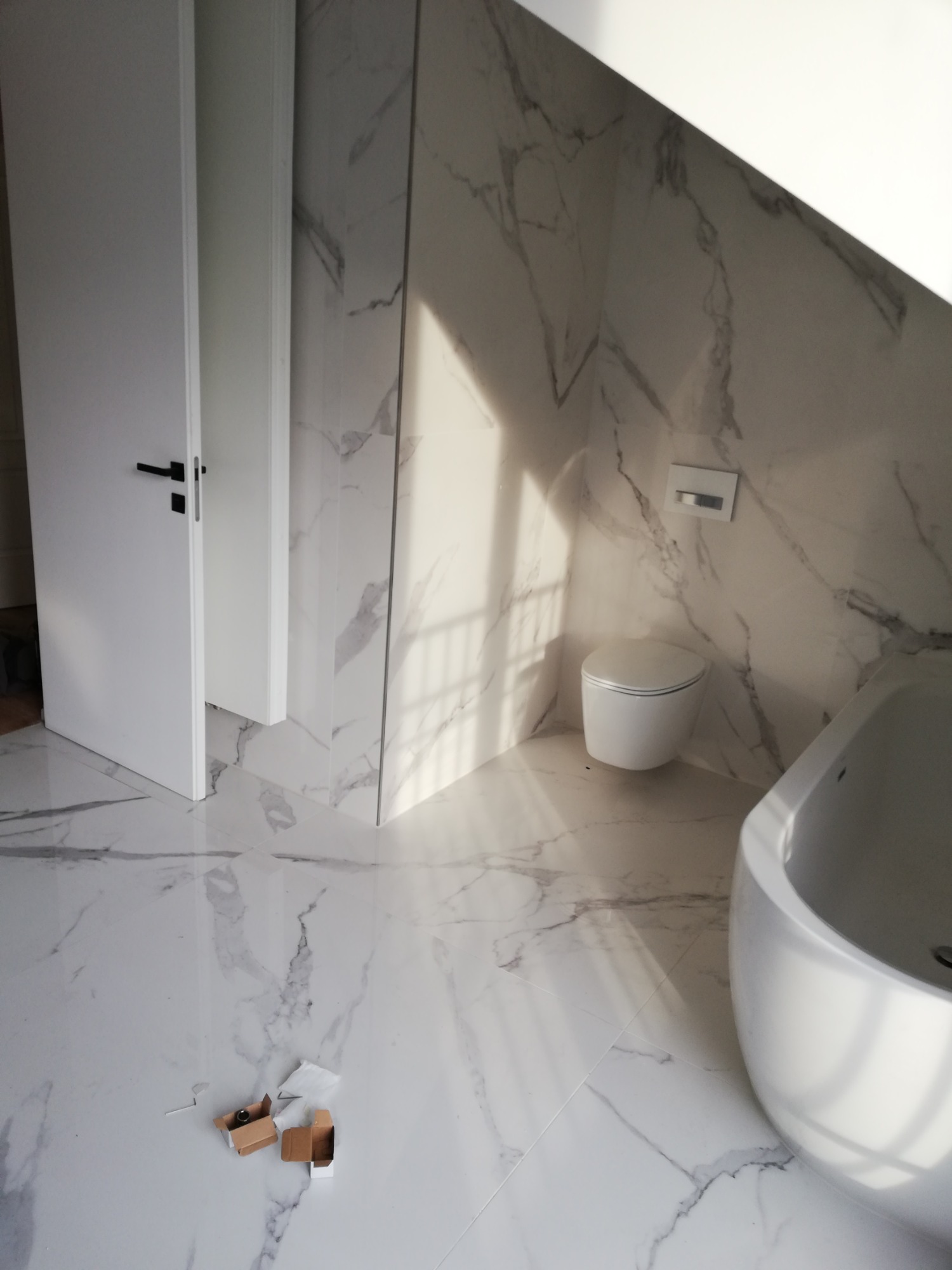 Nowoczesna łazienka z białymi płytkami imitującymi marmur, wisząca toaleta, wolnostojąca wanna i otwarte białe drzwi, z pudełkami po akcesoriach na podłodze.