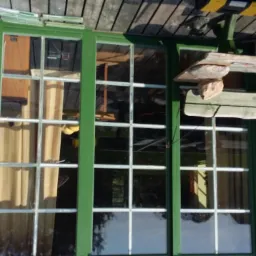 Trzyczęściowe okno z zieloną ramą i białymi szprosami, widok z zewnątrz, z widocznym fragmentem drewnianej werandy i dekoracjami ściennymi.