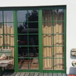 Zielone, przeszklone drzwi tarasowe z widocznymi refleksami otoczenia, flankowane przez kinkiety na białej ścianie, z fragmentem drewnianego tarasu i ławką z akcesoriami na pierwszym planie.