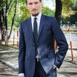 Mężczyzna w granatowym garniturze w prążki, stojący na chodniku w otoczeniu drzew, z ręką w kieszeni i lekkim uśmiechem.