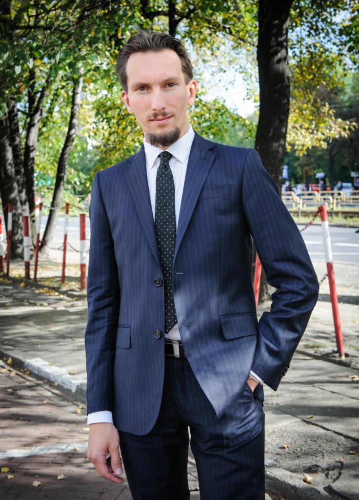 Mężczyzna w granatowym garniturze w prążki, stojący na chodniku w otoczeniu drzew, z ręką w kieszeni i lekkim uśmiechem.