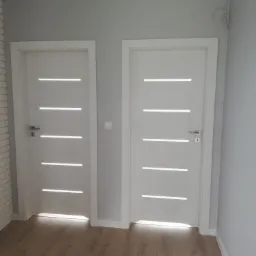 Dwa białe, nowoczesne drzwi wewnętrzne z poziomymi przeszkleniami, zamontowane w szarej ścianie, widoczne na tle paneli podłogowych.