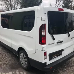 Biały Renault Trafic z przyciemnionymi szybami, widok z tyłu na żwirowym podłożu, z naklejką informacyjną na zderzaku.