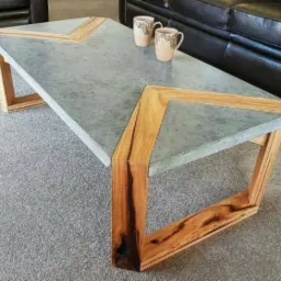 Stolik kawowy z blatem z betonu architektonicznego i drewnianą ramą o geometrycznym wzorze, dwie filiżanki na blacie, widoczna czarna skórzana kanapa w tle.