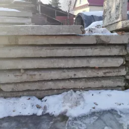 Stos szarych, betonowych płyt ułożonych na sobie w zimowym otoczeniu, częściowo przykrytych śniegiem, z widocznymi fragmentami drewnianych podpór.