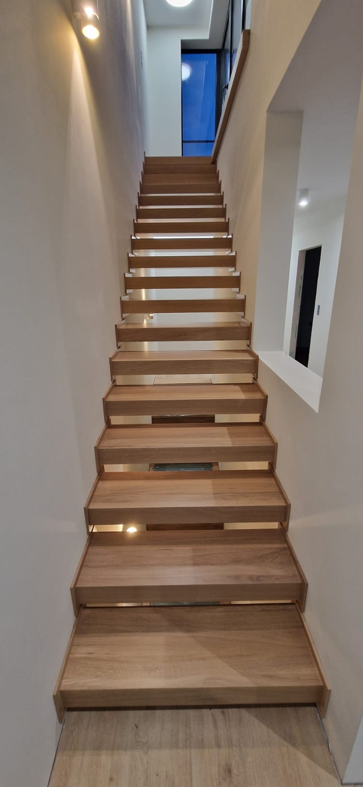 Nowoczesne, drewniane schody wspornikowe z podświetleniem, prowadzące do góry. Minimalistyczny design, jasne ściany, widok z dołu na górę.