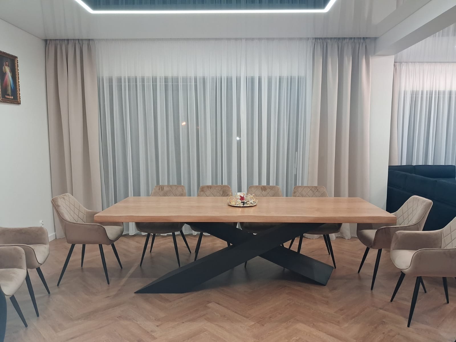 Stół z litego drewna z czarną, krzyżową podstawą w nowoczesnej jadalni. Krzesła tapicerowane w odcieniach beżu, parkiet w jodełkę, jasne zasłony i minimalistyczne oświetlenie.