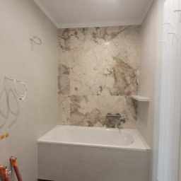 Wykończona łazienka z wanną w zabudowie, marmurowe płytki na ścianie, grzejnik drabinkowy. Widoczne przygotowania do dalszych prac.