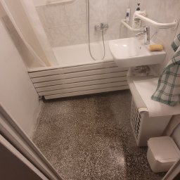 Łazienka z wanną, umywalką i grzejnikiem drabinkowym. Widoczne uchwyty dla niepełnosprawnych. Zdjęcie 'przed' remontem, z widoczną starą podłogą.