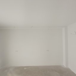 Wnętrze po malowaniu: białe ściany, widoczne gniazdka elektryczne i wystające kable ze ściany. Podłoga przygotowana do dalszych prac wykończeniowych.