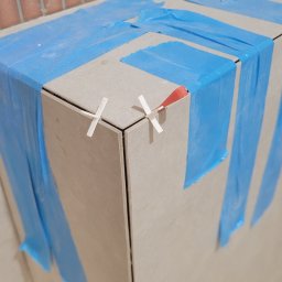 RoKa Renovation Robert Karpiński - Układanie płytek na narożniku, zabezpieczone niebieską folią i krzyżykami dystansowymi. Widoczna ściana z cegieł.