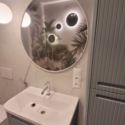RoKa Renovation Robert Karpiński - Nowoczesna łazienka z okrągłym lustrem z podświetleniem LED, umywalką w szafce z szarymi lamelami i marmurową ścianą. Widoczne gniazdka elektryczne.